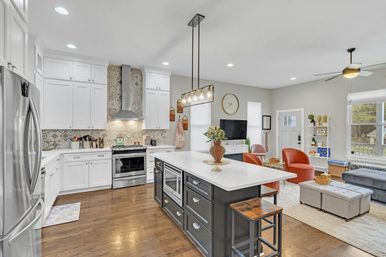 Bright open-concept kitchen and living room with white shaker cabinets, patterned tile backsplash, stainless steel appliances, dark island with white quartz countertop and pendant lights, hardwood floors and cozy seating with orange accent chairs and large windows