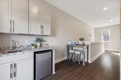 Bright modern kitchenette with white cabinets, marble-look granite countertop and sink, stainless mini fridge, and adjacent rustic folding table with two stools and colorful folded towels on dark hardwood floors.
