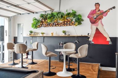 Nashville-themed lounge with illuminated marquee sign amid tropical greenery, retro musician cutout holding a guitar, round white table and beige bar stools on a wood floor with a pool table corner visible.