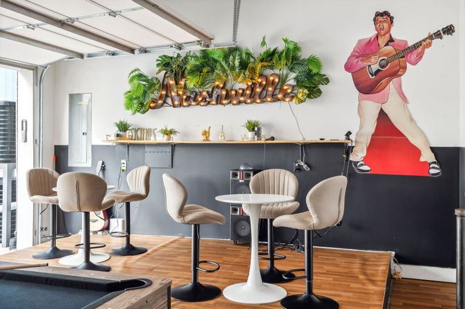 Nashville-themed lounge with illuminated marquee sign amid tropical greenery, retro musician cutout holding a guitar, round white table and beige bar stools on a wood floor with a pool table corner visible.
