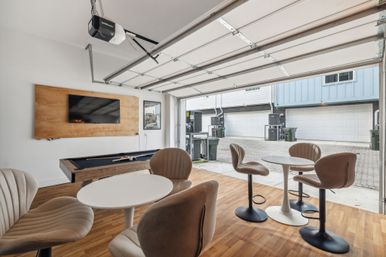 Bright modern garage lounge with open overhead door to paved townhome garages, wood floors, wall-mounted TV, pool table, and round bar-height tables with swivel chairs.