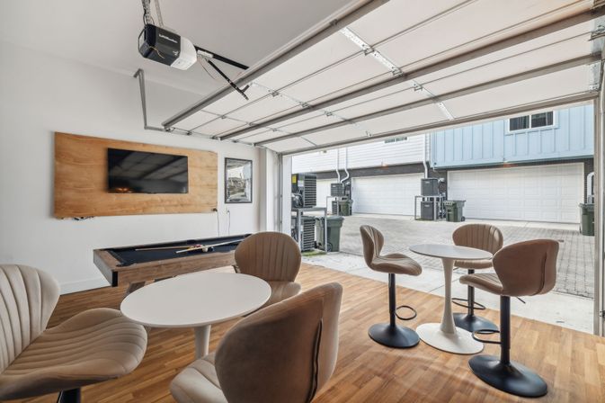 Bright modern garage lounge with open overhead door to paved townhome garages, wood floors, wall-mounted TV, pool table, and round bar-height tables with swivel chairs.