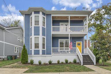 Blue two-story suburban house with white trim and bay windows, second-story balcony and front porch with a yellow door, American flag, stairway, and small landscaped lawn