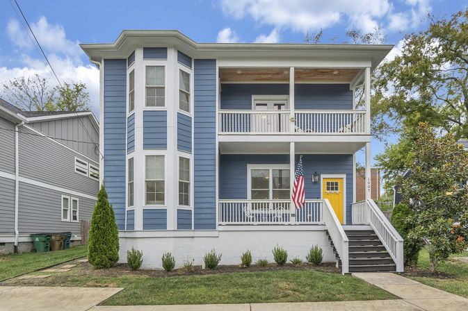 Blue two-story suburban house with white trim and bay windows, second-story balcony and front porch with a yellow door, American flag, stairway, and small landscaped lawn