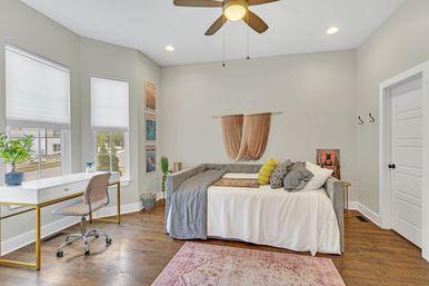 Sunlit modern bedroom with a gray tufted daybed and colorful pillows, macramé wall hanging, white gold‑leg desk by two windows, hardwood floors and ceiling fan — cozy home office nook