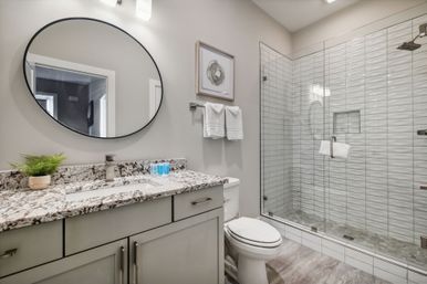 Bright, spa-like bathroom interior with granite countertop vanity, round black-framed mirror, potted plant and toiletries, toilet, and glass-enclosed walk-in shower with textured white tiles.