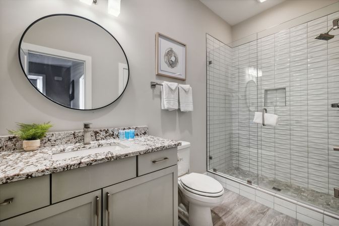 Bright, spa-like bathroom interior with granite countertop vanity, round black-framed mirror, potted plant and toiletries, toilet, and glass-enclosed walk-in shower with textured white tiles.