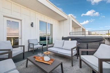 Rooftop patio with modern gray-cushioned lounge chairs and sofa, coffee table with glasses and ice jar, and covered hot tub beside white-paneled building under a bright blue sky.