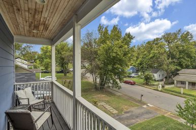 Cozy second-story covered porch with cushioned chairs and white railing overlooking a tree-lined suburban street with houses, lawns, and a blue sky