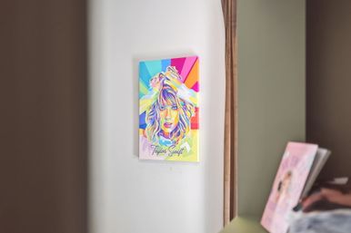 Vibrant pop-art portrait poster of a blonde woman with hands in her hair mounted on a white bedroom wall, reflected in a nearby mirror beside a dresser with books — colorful modern wall art in a cozy home interior.