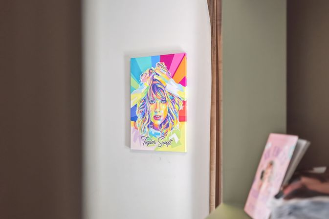 Vibrant pop-art portrait poster of a blonde woman with hands in her hair mounted on a white bedroom wall, reflected in a nearby mirror beside a dresser with books — colorful modern wall art in a cozy home interior.