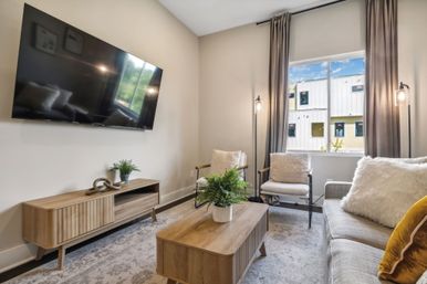 Cozy modern living room with wall-mounted TV, mid-century wooden media console and coffee table, light-gray sofa with textured pillows, two armchairs by a curtained window flooding natural light, and potted green plants in a neutral, airy space.