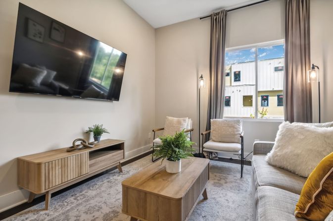 Cozy modern living room with wall-mounted TV, mid-century wooden media console and coffee table, light-gray sofa with textured pillows, two armchairs by a curtained window flooding natural light, and potted green plants in a neutral, airy space.