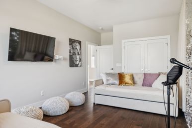 Cozy modern lounge/guest room with a white daybed piled with velvet and metallic throw pillows, wall-mounted flat-screen TV, three knitted poufs on dark hardwood floors, and a microphone on a stand by sequined curtains — ready for a casual hangout or karaoke night.