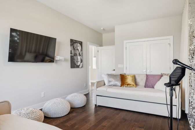 Cozy modern lounge/guest room with a white daybed piled with velvet and metallic throw pillows, wall-mounted flat-screen TV, three knitted poufs on dark hardwood floors, and a microphone on a stand by sequined curtains — ready for a casual hangout or karaoke night.