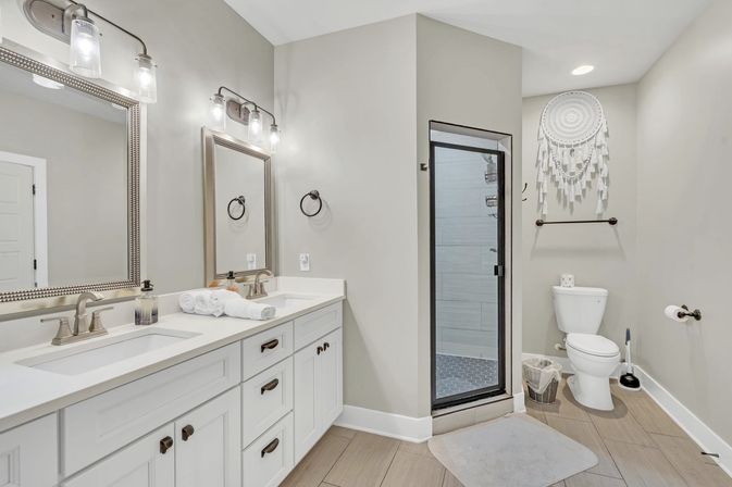 Bright modern master bathroom with white double vanity and quartz countertop, two framed mirrors and brushed nickel faucets, glass-door shower with gray tile, toilet and boho macrame wall hanging on neutral beige walls over wood-look floor.