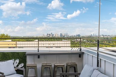 Sunny rooftop patio with cushioned lounge seating, metal bar stools and railing overlooking a distant downtown skyline and tree-lined cityscape.