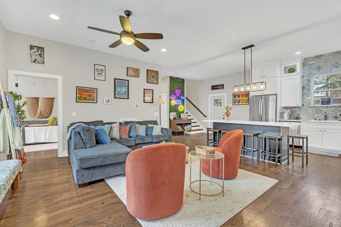 Modern open-concept living room and kitchen in a suburban home with hardwood floors, gray sectional, two coral swivel chairs, gold side table, island with stools, stainless fridge, patterned backsplash and neon wall art by the stairs.