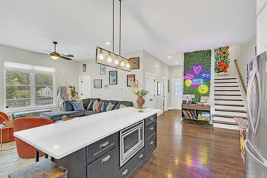 Bright open-concept living area with white kitchen island and industrial pendant lights, dark cabinets with built-in microwave, gray sectional and orange chairs, hardwood floors, staircase, and a green accent wall with colorful neon signs.