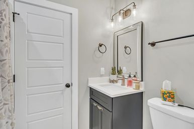 Small modern bathroom with white paneled door, neutral gray walls, dark gray vanity with white countertop and sink, framed mirror under a three-bulb sconce, potted succulent and soap dispensers on the counter, patterned shower curtain and toilet with colorful tissue box.