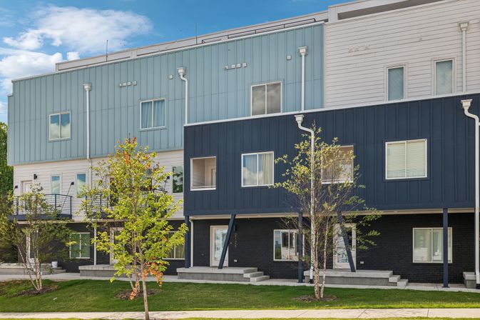 Contemporary blue-and-white multi-level townhouses with metal siding, front steps, lawns and young trees in a suburban neighborhood.