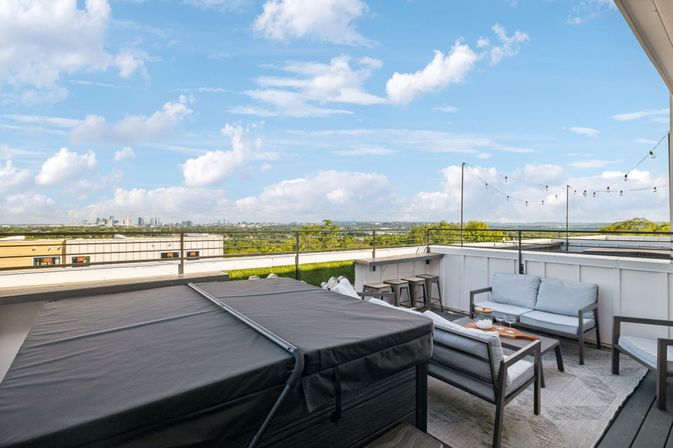 Rooftop patio lounge with a covered hot tub, cushioned sofas, coffee table and bar stools under string lights, overlooking a distant downtown skyline and bright blue sky with clouds.