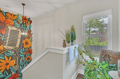 Bright home stair landing with a colorful mural of a vintage microphone and oversized orange flowers, geometric pendant light, white half-wall topped with a blue runner and potted grasses, window overlooking a green yard, patterned chair and lush indoor plants.