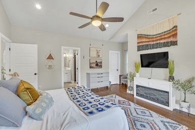Bright contemporary master bedroom in a suburban home with vaulted ceiling and ceiling fan, white bedding with blue patterned throw, hardwood floors, wall-mounted TV above a white fireplace, woven wall hanging, gray dresser, potted plants and en-suite bathroom visible.
