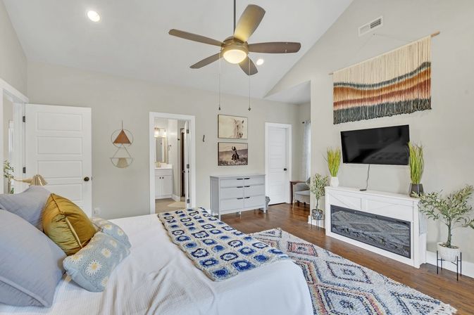 Bright contemporary master bedroom in a suburban home with vaulted ceiling and ceiling fan, white bedding with blue patterned throw, hardwood floors, wall-mounted TV above a white fireplace, woven wall hanging, gray dresser, potted plants and en-suite bathroom visible.