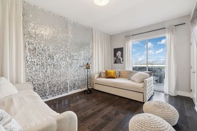 Bright modern condo living room with sparkling silver sequin accent wall, cream tufted daybeds with velvet and faux-fur pillows, knitted poufs, dark hardwood floors, a floor lamp, and a sliding glass door opening to a balcony under a blue sky.