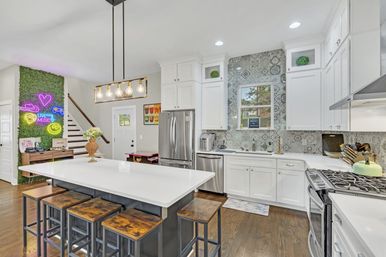 Bright modern open-concept residential kitchen with large white island on a dark base, four rustic wooden stools, white shaker cabinets, patterned gray tile backsplash, stainless-steel appliances and gas range, Edison-bulb pendant light, hardwood floors, and a green moss accent wall with neon art by the staircase.