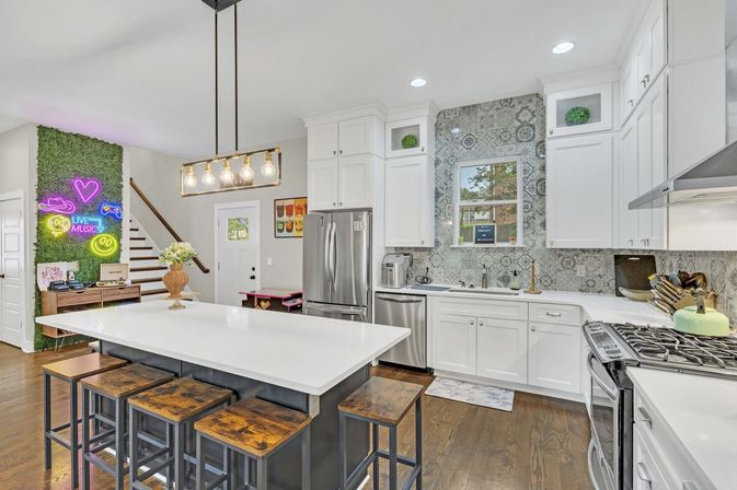 Bright modern open-concept residential kitchen with large white island on a dark base, four rustic wooden stools, white shaker cabinets, patterned gray tile backsplash, stainless-steel appliances and gas range, Edison-bulb pendant light, hardwood floors, and a green moss accent wall with neon art by the staircase.