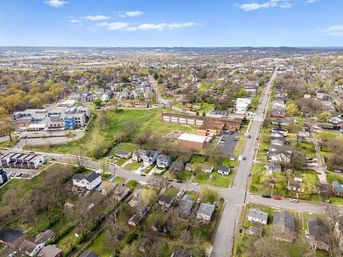 Aerial view of a suburban neighborhood with tree-lined streets, single-family homes, a school complex, parking lots and open green spaces under a blue sky.