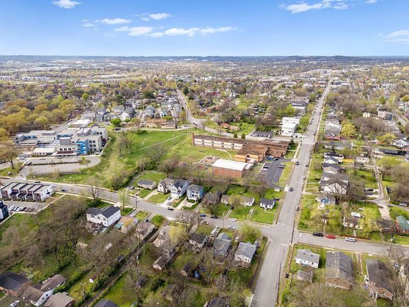 Aerial view of a suburban neighborhood with tree-lined streets, single-family homes, a school complex, parking lots and open green spaces under a blue sky.