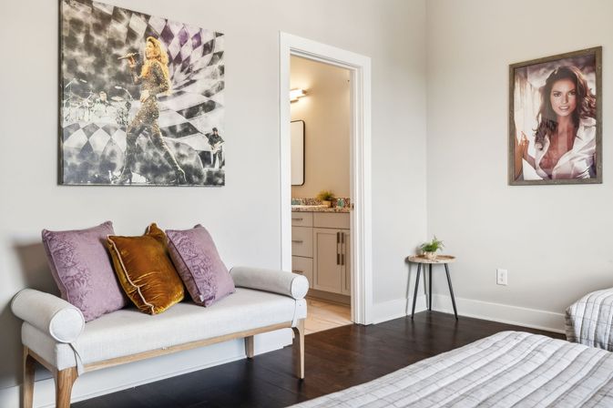 Modern bedroom interior with dark hardwood floors, light upholstered bench topped with purple and gold pillows, framed wall art, doorway to an ensuite bathroom, and a small round side table with a potted plant.
