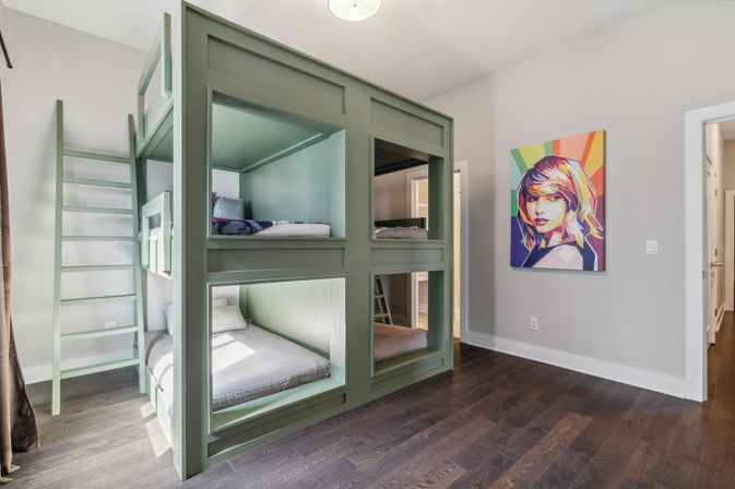 Sunlit modern kids' room with a built-in green four-cubby bunk bed and ladder, dark hardwood floors, and a colorful pop-art portrait on a light gray wall.