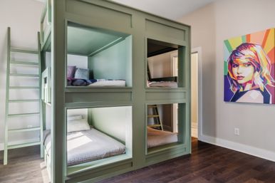 Modern kids' bedroom with sage-green built-in four-person bunk beds and ladders, cozy mattresses, dark hardwood floors and a colorful pop-art portrait on the wall.