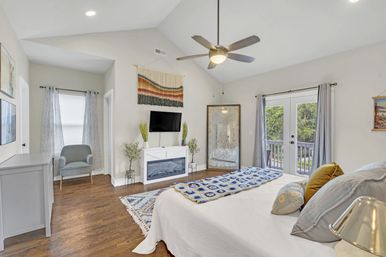Sunlit, cozy modern master bedroom with vaulted ceiling and ceiling fan, hardwood floors, white bed topped with blue crocheted throw and pillows, wall-mounted TV above a white electric fireplace flanked by plants, woven wall hanging, full-length mirror, armchair, patterned rug, and French doors opening to a balcony.