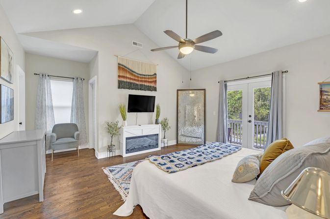 Sunlit, cozy modern master bedroom with vaulted ceiling and ceiling fan, hardwood floors, white bed topped with blue crocheted throw and pillows, wall-mounted TV above a white electric fireplace flanked by plants, woven wall hanging, full-length mirror, armchair, patterned rug, and French doors opening to a balcony.