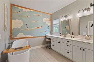 Modern residential bathroom with freestanding white tub and wooden caddy, long white double vanity with two mirrors and lights, neutral tile floor, and a large framed whimsical mural of swimmers flying through clouds.