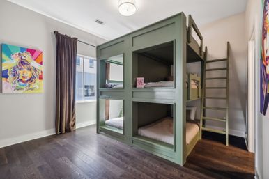 Kids' bedroom with sage-green cube-style bunk beds (four sleeping cubbies) and ladder, dark hardwood floors, a window with brown curtains letting in daylight, and a vibrant pop-art portrait poster on the wall.