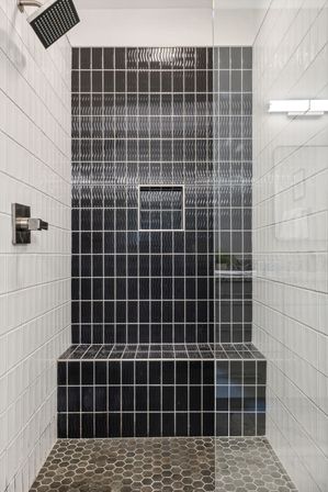 Sleek modern bathroom shower with glass enclosure, hexagon stone floor, white square wall tiles, a floor-to-ceiling black wavy-tile accent wall with built-in niche and bench, and a square rain showerhead.