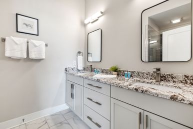 Spa-like modern bathroom interior with double-sink granite countertop, light-gray shaker cabinets, rectangular wall mirrors, white folded towels on a rack, marble-look floor tiles, and a small potted plant.