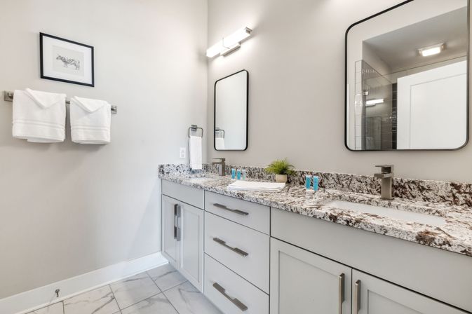 Spa-like modern bathroom interior with double-sink granite countertop, light-gray shaker cabinets, rectangular wall mirrors, white folded towels on a rack, marble-look floor tiles, and a small potted plant.