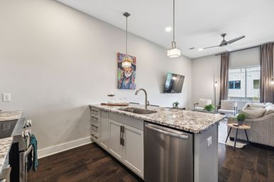 Open-concept modern kitchen and living area with speckled granite island and sink, stainless steel dishwasher, pendant lights, dark hardwood floors, wall-mounted TV and neutral sofa seating by a large window.