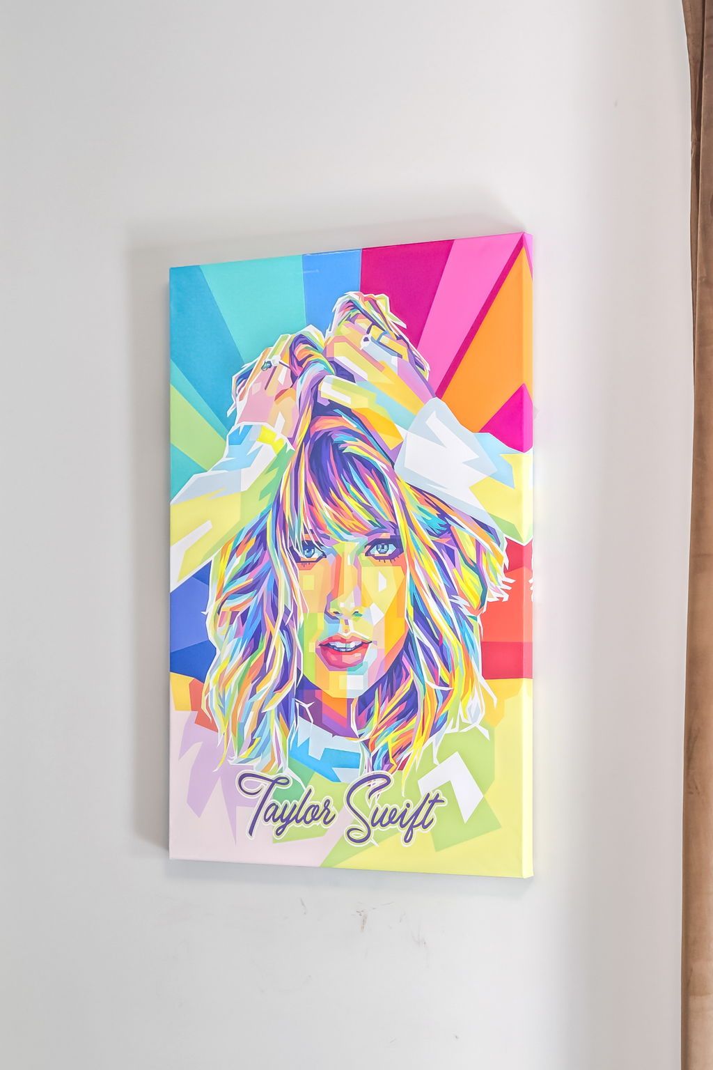 Vibrant pop-art canvas portrait of a female pop singer in rainbow colors with hands in her hair and geometric rays, hung on a white wall next to a beige curtain.