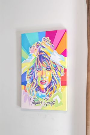 Vibrant pop-art canvas portrait of a female pop singer in rainbow colors with hands in her hair and geometric rays, hung on a white wall next to a beige curtain.