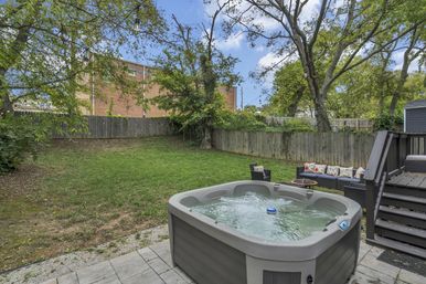 Cozy residential backyard with a bubbling hot tub on a stone patio, outdoor sofa and chairs with colorful cushions, grassy lawn, tall wooden privacy fence, mature trees and hanging string lights.