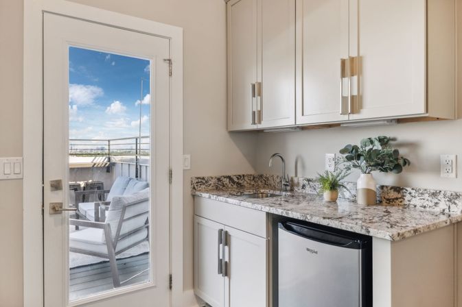 Sunny modern kitchenette with granite countertop, sink and stainless mini fridge, glass door leading to rooftop patio with cushioned outdoor chairs and blue sky