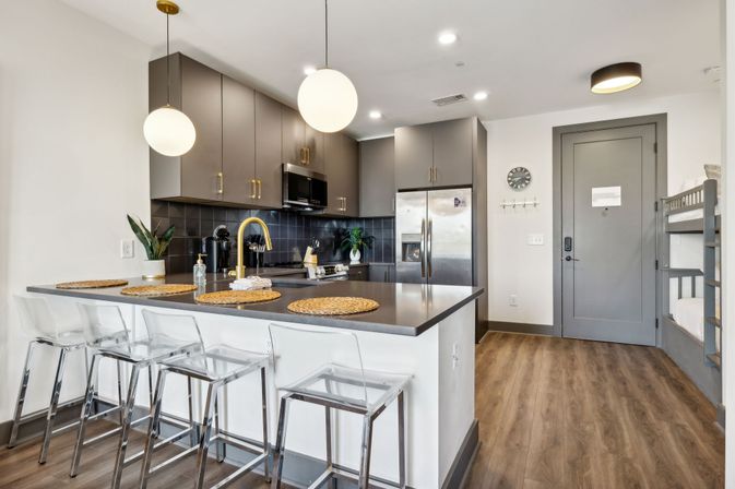 Open-concept modern apartment kitchen with gray cabinets, stainless steel fridge, dark tile backsplash, island breakfast bar with clear stools and gold faucet, wood floors and bunk beds by the entry